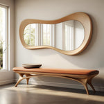 May include: A modern, organic-shaped mirror with a light wood frame hangs above a bench. The bench has a brown leather seat and light wood legs. A small, round, gray bowl sits on the bench.