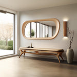 May include: A light wood bench with a curved, organic design and a beige cushion. The bench is in front of a large, oval mirror with a wood frame. A wall-mounted light fixture is on the right side of the mirror.