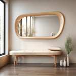 May include: A light wood bench with a white cushion and a large oval mirror with a light wood frame hanging above it. There are two white vases and a small wooden vase on the floor next to the bench.
