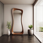 May include: A large, modern, wooden framed mirror with a wavy, organic shape. The mirror is standing in a room with white walls and a large window. There are two potted plants on either side of the mirror.