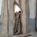 May include: A full-length mirror with a unique, organic, wavy wooden frame. The frame is a dark brown color and the mirror reflects a stack of wooden planks.