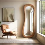 May include: A full-length wooden mirror with a unique, organic shape. The mirror is standing in a room with a beige wall and a brown leather armchair.