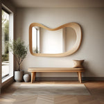 May include: A wooden bench with a light brown cushion sits in front of a large, wavy, wooden framed mirror. A small potted plant is to the left of the bench.