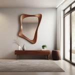 May include: A modern living room with a wooden console table, a large, abstract, wooden framed mirror, a potted plant, and a brown pouf. The console table has three drawers and sits on tapered legs. The mirror is mounted on the wall behind the console table. The pouf is placed to the right of the console table. The room has a large window with white curtains.