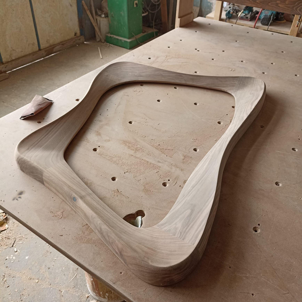 May include: A light-colored, unfinished wooden frame with a unique, curved design. The frame has rounded corners and a smooth surface, suggesting it's for a mirror or artwork. A piece of sandpaper rests nearby.