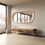 May include: A light wood bench with a curved, organic design and a beige cushion. The bench is in front of a large, oval-shaped mirror with a light wood frame. The mirror reflects a room with a window and a flowering tree.