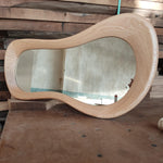 May include: A wooden mirror with a unique, wavy, organic shape. The mirror is made of light-colored wood and has a smooth, polished finish.