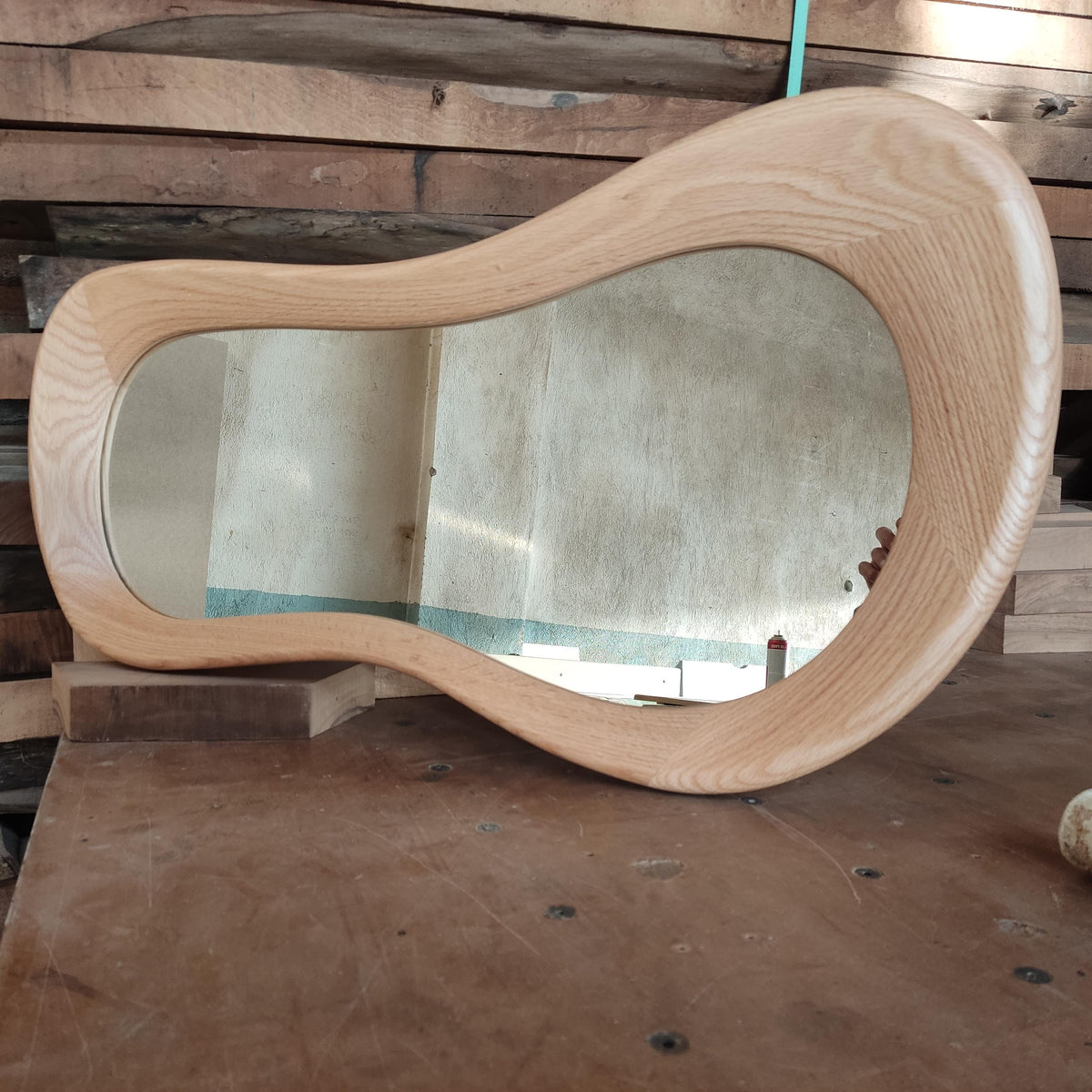 May include: A wooden mirror with a unique, wavy, organic shape. The mirror is made of light-colored wood and has a smooth, polished finish.