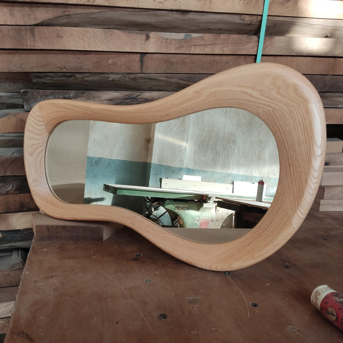 May include: A wooden mirror with a unique, wavy, organic shape. The mirror is made of light-colored wood and has a reflective surface.