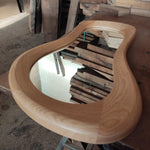 May include: A wooden mirror with a unique, organic shape. The mirror has a natural wood finish and a smooth, rounded edge. The mirror is reflecting a stack of wooden planks.