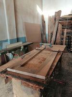 May include: A wooden table top is being assembled with clamps on a workbench. The table top is made of light brown wood with a natural grain pattern. The workbench is made of dark wood and has a worn surface. There are various pieces of wood stacked in the background.