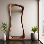 May include: A large, free-form wooden mirror with a brown finish. The mirror is standing on a dark floor in front of a white wall. There are two potted plants on either side of the mirror.