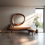 May include: A wooden console table with a unique, organic shape and a large, oval mirror above it. The table has a light wood finish and is supported by two legs on each side. A small, round ottoman with a light-colored cushion sits in front of the table. The room has a minimalist design with a white wall and a large window.