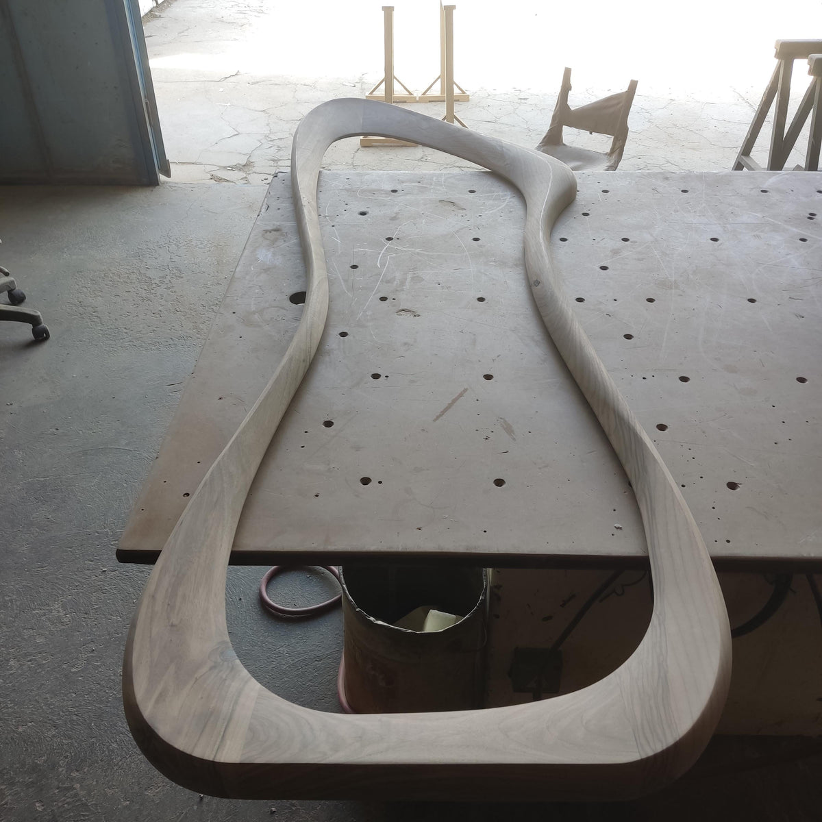 May include: A large, unfinished, wooden frame with a curved, organic shape. The frame is resting on a gray surface.