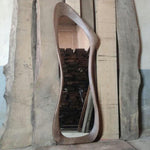 May include: A large, free-form wooden mirror with a natural wood finish. The mirror is leaning against a stack of wooden planks.
