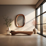 May include: A wooden bench with a curved design sits in a room with a large window and a wooden oval mirror on the wall.  A small side table with a vase of greenery sits to the left of the bench.