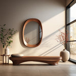 May include: A wooden bench with a curved design and a cushioned seat sits in front of a large oval mirror with a wooden frame. A small table with a vase of greenery sits to the left of the bench, and a vase with a leafy branch sits to the right. The room has a light beige wall and a large window with a view of trees.
