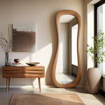 May include: A large, free-form wooden mirror with a light wood finish. The mirror is standing in a room with a wooden dresser and a potted plant.