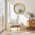 May include: A white armchair with wooden legs and arms sits in a room with a large round mirror with a wooden frame and a wooden dresser with two drawers. The room is decorated in a minimalist style with natural light.