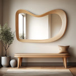 May include: A wooden bench with a cushioned seat and a large, organic-shaped mirror above it. The mirror has a light wood frame and reflects a window with a view of trees.