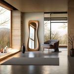 May include: A modern, wooden framed full-length mirror with a curved design, standing in a room with a large window and a gray armchair.