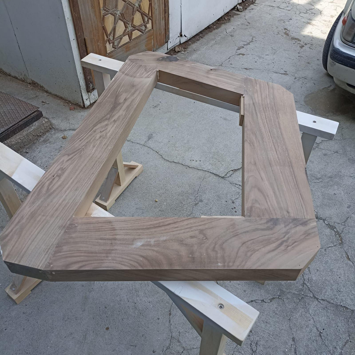 May include: A rectangular wooden frame with a light brown finish. The frame is constructed from thick planks of wood, with a large rectangular opening in the center. The wood grain is visible, and the frame is resting on two sawhorses.
