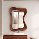 May include: A wavy-edged, brown wooden mirror hangs above a bathroom vanity. The mirror's frame has a unique, organic shape. The vanity features a white countertop, a white oval sink, and a dark brown wooden cabinet.