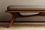 May include: A modern bench with a brown wooden frame and a gray upholstered seat. The bench has a curved design with a smooth, polished finish. The legs are angled and provide a sturdy base. The overall design is minimalist and elegant.