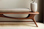 May include: A modern wooden bench with a curved design and a light-colored cushion. A decorative vase with dried stems sits on the bench. The bench is made of dark wood and has a unique, sculptural form.