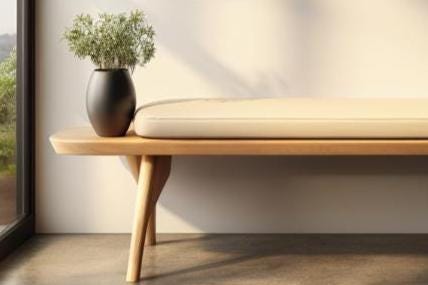 May include: A wooden bench with a light-colored cushion and angled legs. A black vase with greenery sits on the bench. The bench is in front of a window and a white wall.