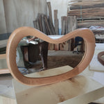 May include: A unique, heart-shaped mirror with a light brown wooden frame. The mirror is displayed on a light-colored wooden surface, showcasing its smooth, curved design. The frame's natural wood grain adds to its aesthetic appeal.