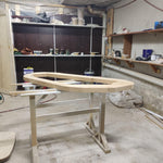 May include: A light-colored, unfinished wooden boat frame sits on a white and light-colored wooden sawhorse. The frame is oval-shaped with a flat top and angled sides. The workshop background includes shelves, tools, and a dark brown cabinet.