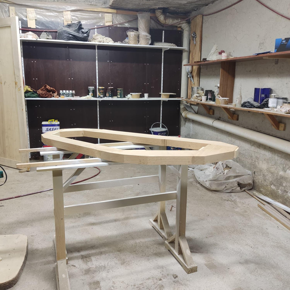 May include: A light-colored, unfinished wooden boat frame sits on a white and light-colored wooden sawhorse. The frame is oval-shaped with a flat top and angled sides. The workshop background includes shelves, tools, and a dark brown cabinet.