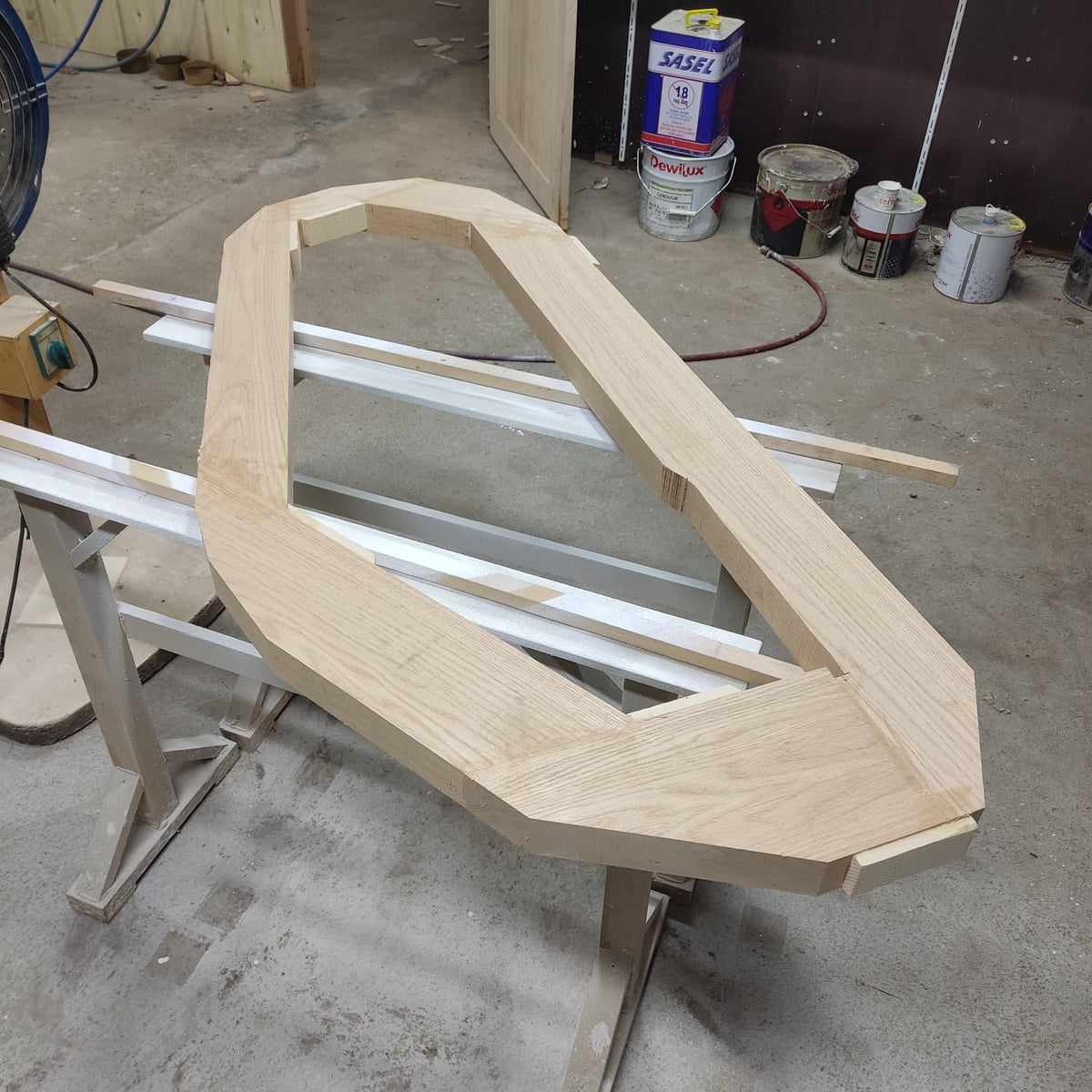 May include: A light-colored, unfinished wooden frame with an arched top and angled sides. The frame is resting on a white sawhorse in a workshop setting. The wood grain is visible.