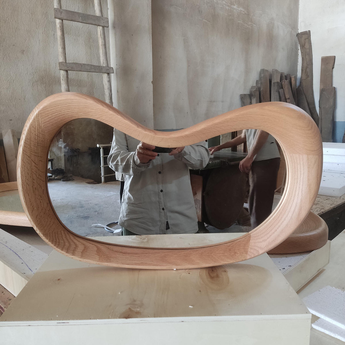 May include: A large, organically shaped wooden mirror. The frame is made of light-colored wood and has a smooth, curved design. The mirror reflects the surrounding workshop environment, including a person holding a phone.