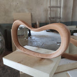May include: A modern, oval-shaped wooden mirror with a light brown finish. The frame has a unique, flowing design. The mirror is resting on a light-colored wooden surface, showcasing its smooth curves and natural wood grain.