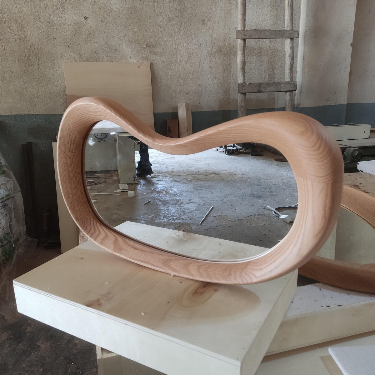 May include: A modern, oval-shaped wooden mirror with a light brown finish. The frame has a unique, flowing design. The mirror is resting on a light-colored wooden surface, showcasing its smooth curves and natural wood grain.
