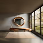 May include: A modern living room featuring a large, oval-shaped wooden mirror and a matching console table. The room has a neutral color palette with a beige rug and a large window overlooking a green landscape. A vase with branches and a potted plant add natural elements.