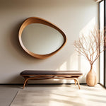 May include: A modern interior scene featuring a large, organically shaped wooden mirror mounted on a neutral wall. Below, a wooden bench with a dark cushion complements the mirror. A tall vase with branches stands near a window, enhancing the room's minimalist aesthetic.