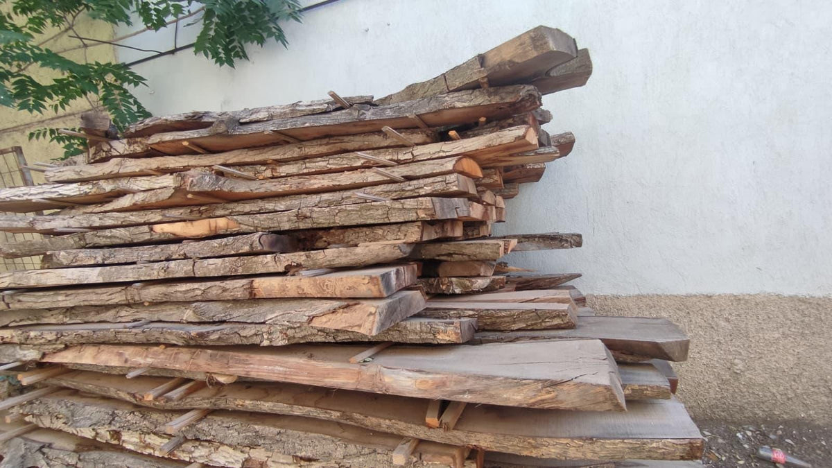 May include: A stack of rough-hewn wooden planks, varying in size and thickness, with natural bark edges. The wood has a warm, brown tone, suggesting it's been weathered. The planks are stacked against a white wall.