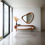 May include: A modern interior featuring a wooden console table with a matching organic-shaped mirror. A dark vase with dried flowers and a small bowl sit on the table. The room has large windows and a minimalist design with a neutral color palette.