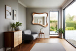 May include: A modern living room with a large, wavy-edged wooden mirror as the focal point. The room features a gray sofa, a wooden cabinet, and framed artwork. A large window offers a view of a green outdoor space. The room has hardwood floors and a light gray rug.