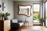 May include: A modern living room with a large, uniquely shaped wooden mirror. A gray sofa, small side table, and wooden cabinet complement the room's neutral color palette. Green plants in vases add a touch of nature. The room has a large window.