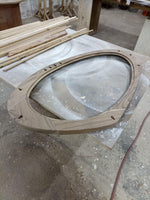 May include: Oval wooden frame with pre-drilled holes and a groove for glass or a mirror. The frame is light brown and appears to be unfinished. Several wooden planks and other woodworking materials are in the background.