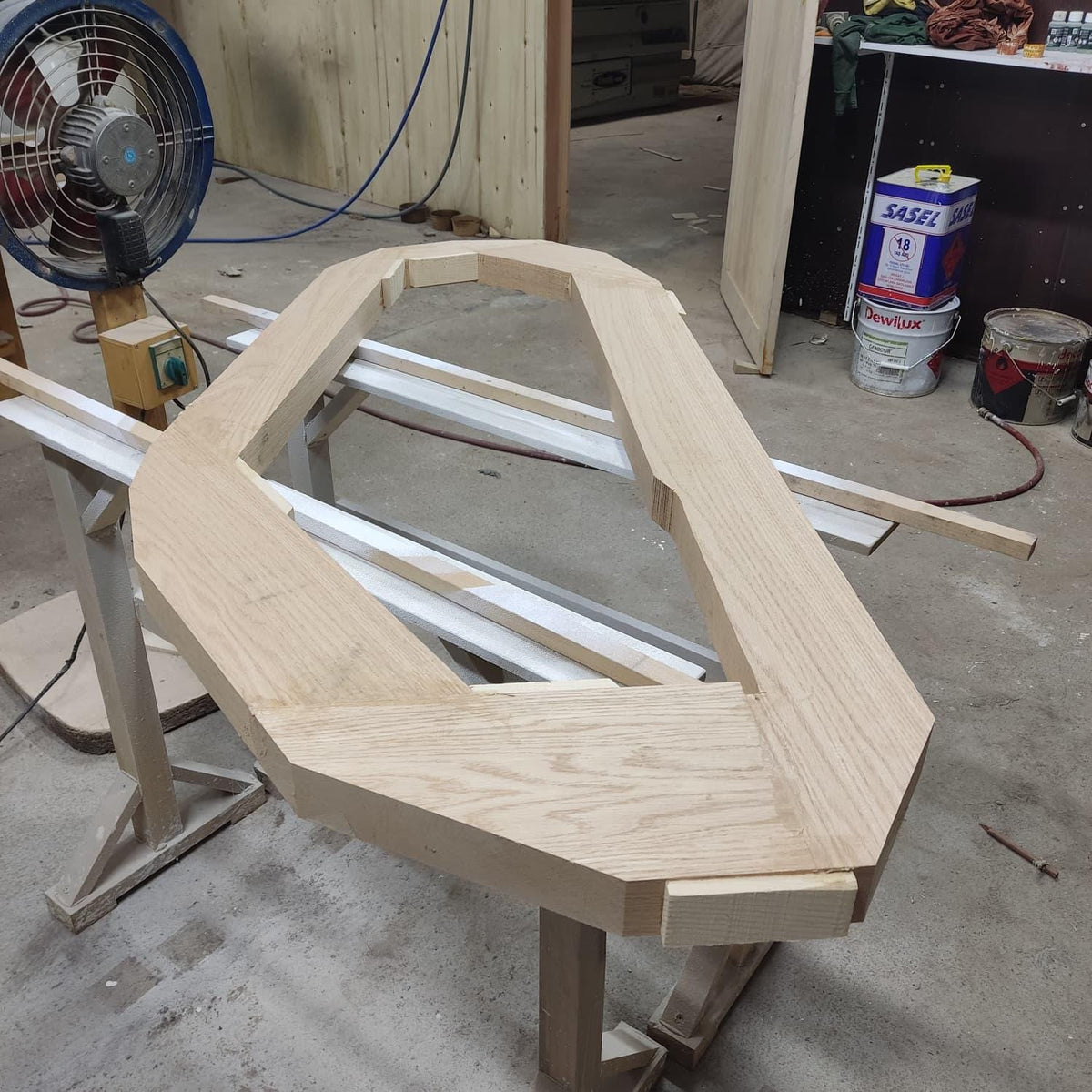 May include: A light-colored, unfinished wooden frame, possibly for a door or window, rests on a sawhorse. The frame has a unique geometric shape with angled corners and a rectangular opening. A fan is visible in the background.