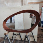 May include: A large, oval-shaped wooden mirror with a dark brown frame. The mirror is supported by a metal sawhorse. The frame has a smooth, curved design, and the mirror reflects the surrounding workshop environment.