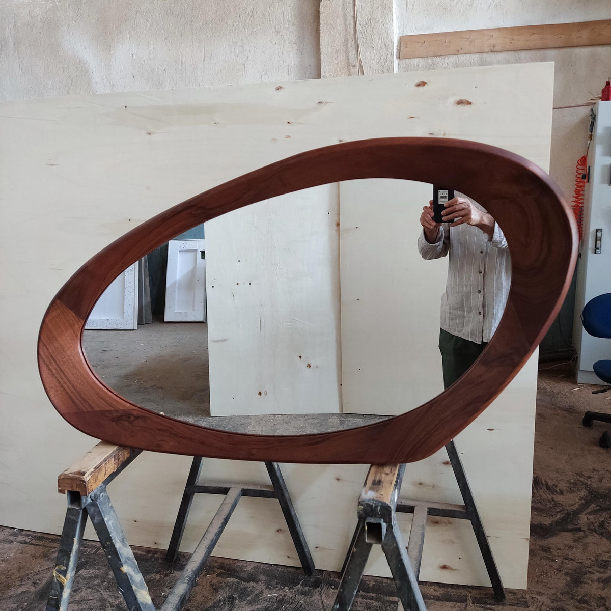 May include: A large, oval-shaped wooden mirror with a dark brown frame. The mirror is supported by a metal sawhorse. The frame has a smooth, curved design, and the mirror reflects the surrounding workshop environment.