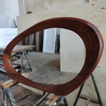 May include: A large, oval-shaped mirror with a dark brown wooden frame. The frame has a smooth, curved design. The mirror is displayed on a wooden sawhorse in a workshop setting. The wood grain is visible.