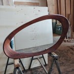 May include: A large, oval-shaped wooden frame with a rich, dark brown finish. The frame is supported by a metal sawhorse in a workshop setting. The wood grain is visible, and the frame appears to be a decorative element.