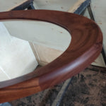 May include: Close-up of a wooden-framed oval mirror. The frame is a rich, dark brown with visible wood grain. The mirror's surface reflects the surrounding environment, creating a sense of depth and dimension. The design is simple and elegant.